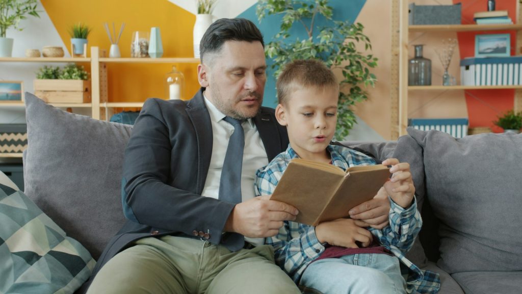 A tutor reading a book together with a young boy on a couch at home