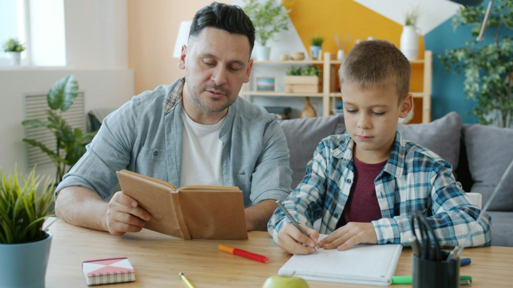 An adult reading aloud while a boy draws at a table at home